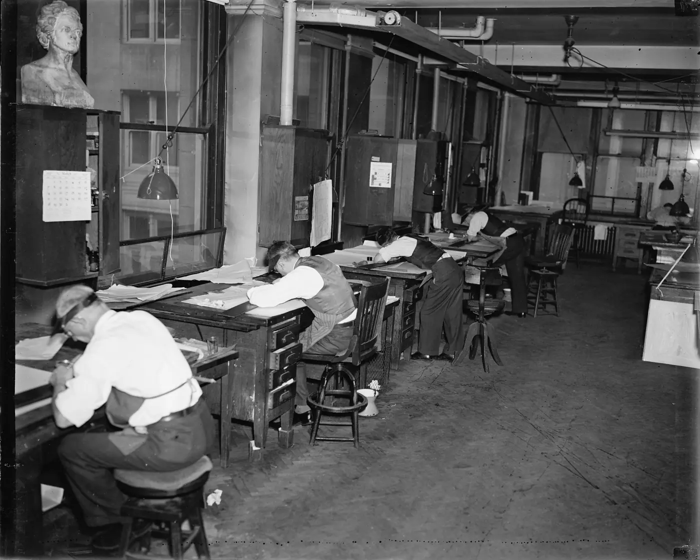Photo de 1937 : des dessinateurs lithographes penchés sur leurs tables, en gilet, corrigent des plaques d'impression de cartes géologiques sous des lampes suspendues.