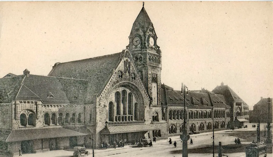 Carte postale ancienne sépia : la gare de Metz, longue façade en pierre avec sa tour d'horloge, une automobile des années 1920 et quelques passants sur la place.