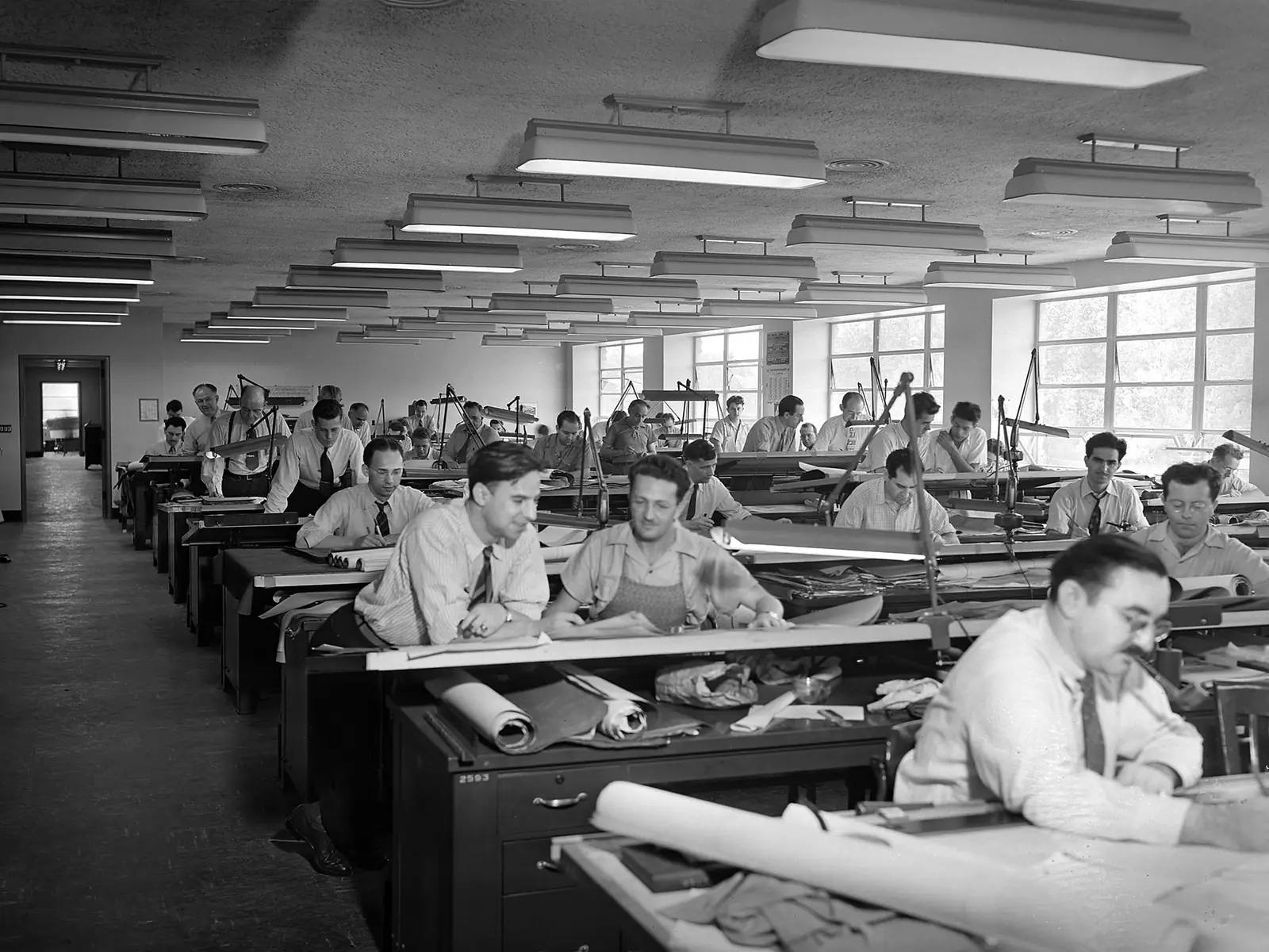 Photo noir et blanc de 1942 : une grande salle de dessin remplie de tables à dessin inclinées, une quarantaine d'hommes en chemise et cravate penchés sur des plans sous des rangées de néons.